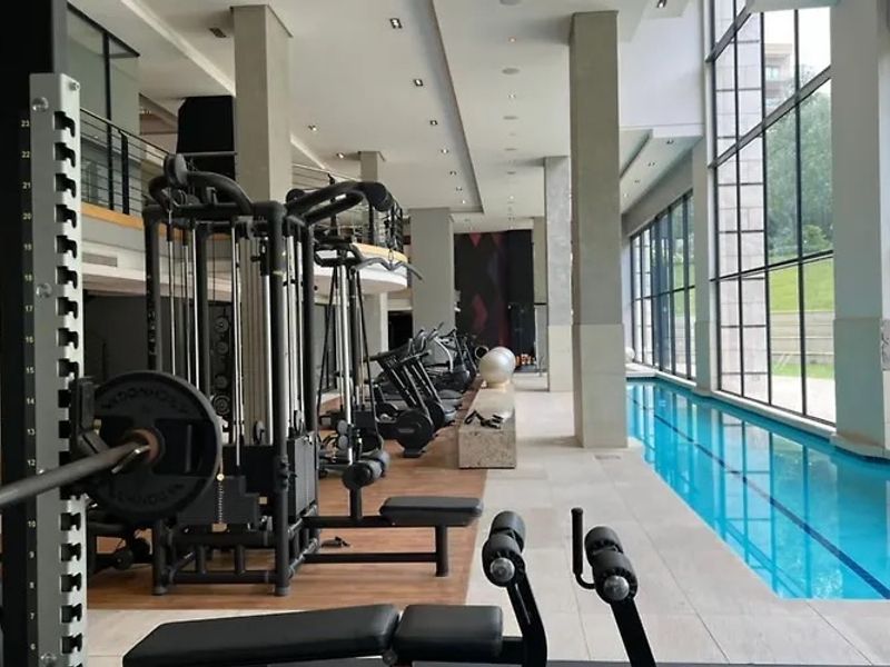 A modern, well-maintained indoor gym facility featuring professional exercise equipment, large floor-to-ceiling windows, and an adjacent indoor lap pool.