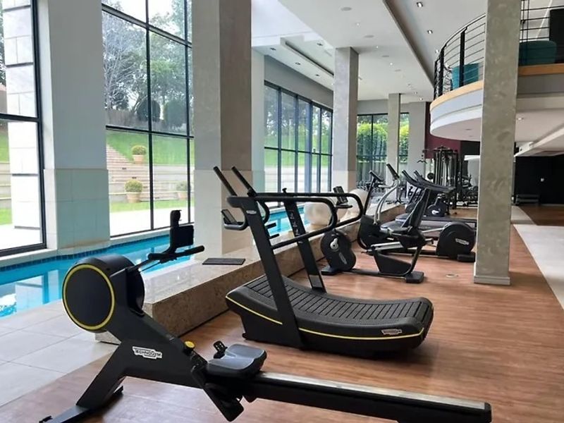 Modern gym facility featuring high ceilings, floor-to-ceiling windows, and an integrated indoor swimming pool with a mezzanine level.
