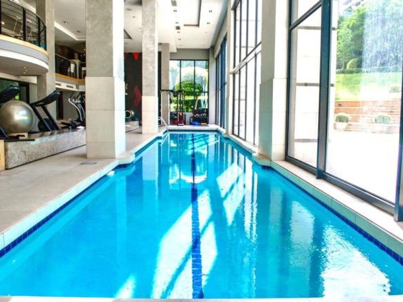 A modern indoor lap pool featuring floor-to-ceiling windows, concrete structural columns, and a clean tiled surround, adjacent to a fitness area.