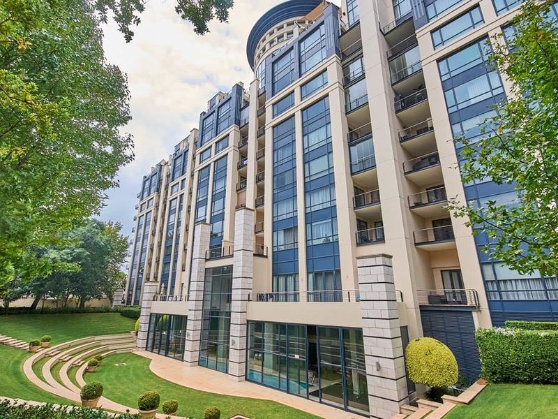 A view of the rear facade of a modern multi-story apartment building featuring glass curtain walls, private balconies, and well-maintained landscaped gardens.