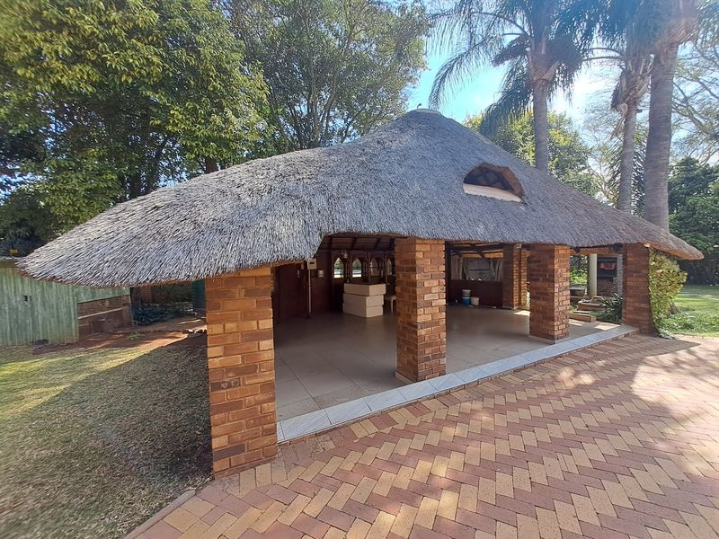 Spacious outdoor entertainment area featuring a traditional thatched roof, brick pillars, tiled flooring, and a built-in braai, set within a garden environment.