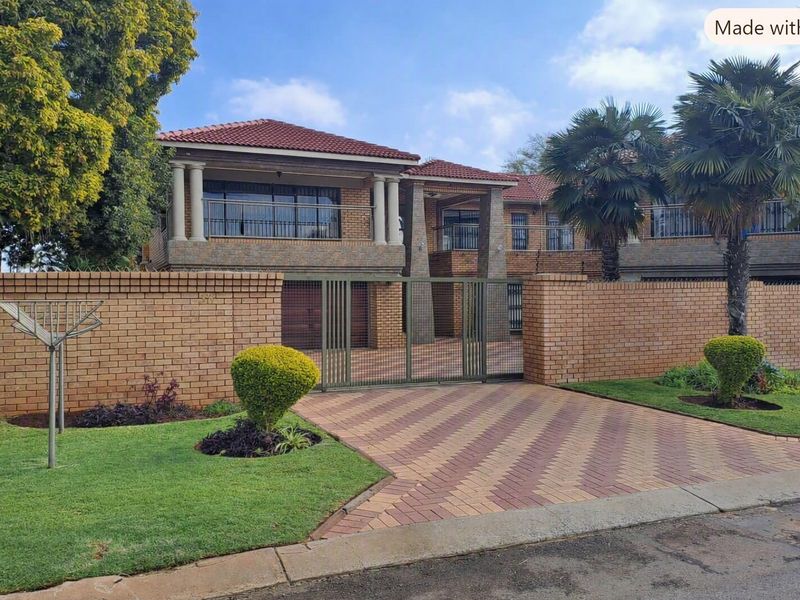 Front view of a double-story brick house featuring a paved driveway, secure metal gate, boundary wall, and upper-level balconies with columns.