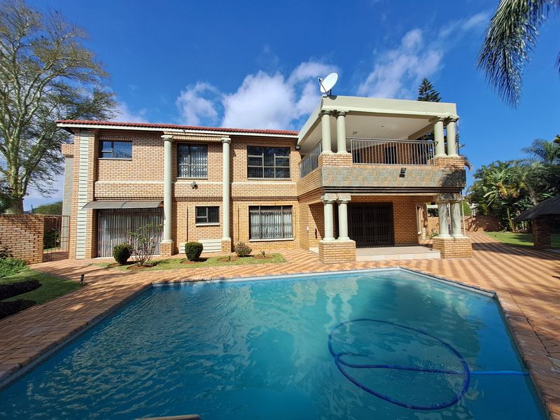 Rear view of a double-story brick house featuring a swimming pool, paved patio area, and an upper-level balcony with decorative columns.