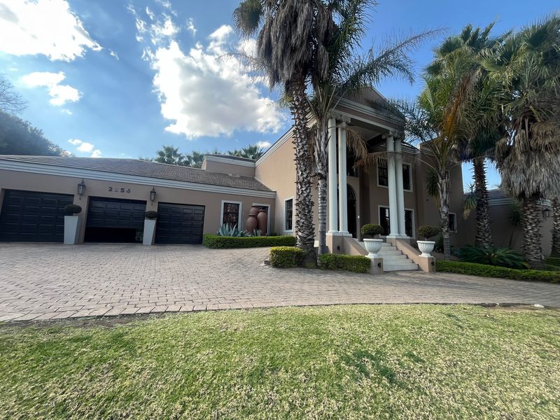 A grand front elevation featuring a paved driveway, double garage, and an impressive portico entrance with columns, set within a well-maintained garden.