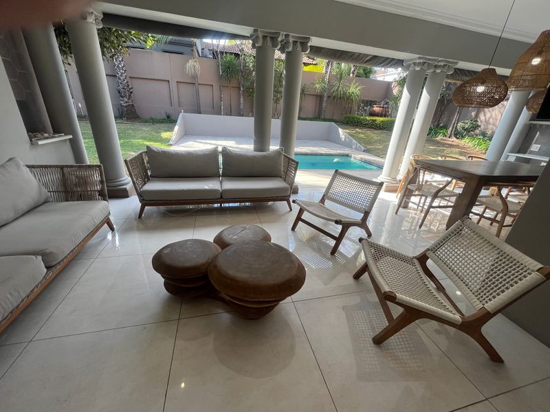 A spacious covered patio featuring tiled flooring and structural columns, overlooking a private swimming pool and landscaped garden area.