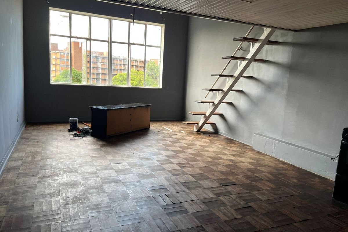 An open-plan living area featuring original parquet flooring, a metal staircase leading to a mezzanine level, and a large window with urban views.