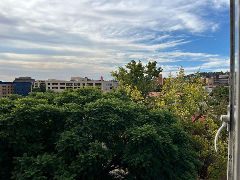 An elevated urban view overlooking mature trees and surrounding apartment buildings under a partly cloudy sky.