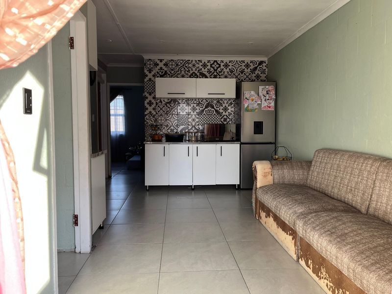 Open plan living area featuring a compact kitchen with modern white cabinetry and a decorative patterned tile backsplash, complemented by neutral floor tiling.