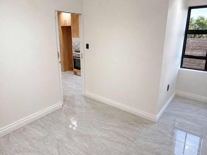 A bright, empty living area featuring modern porcelain tile flooring, crisp white walls, and a view into the adjacent kitchen space.