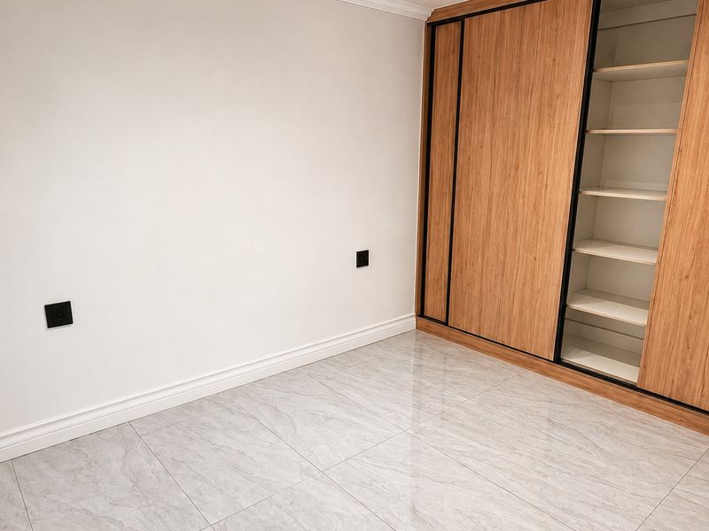A bright, empty bedroom featuring modern large-format marble-look floor tiles, crisp white walls, and a contemporary built-in wooden wardrobe with sliding doors.