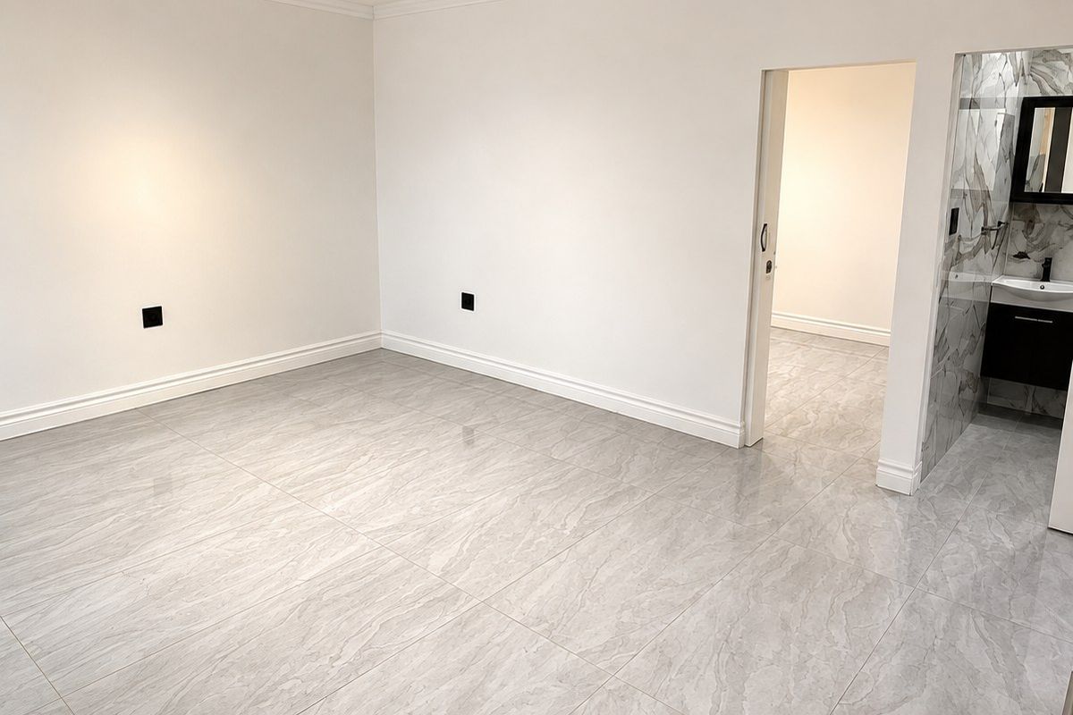 A bright, empty bedroom featuring modern grey porcelain tile flooring, crisp white walls, and an adjoining en-suite bathroom with marble-effect tiling.