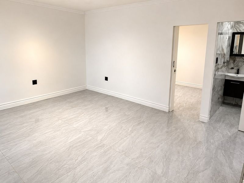 A bright, empty bedroom featuring modern grey porcelain tile flooring, crisp white walls, and an adjoining en-suite bathroom with marble-effect tiling.