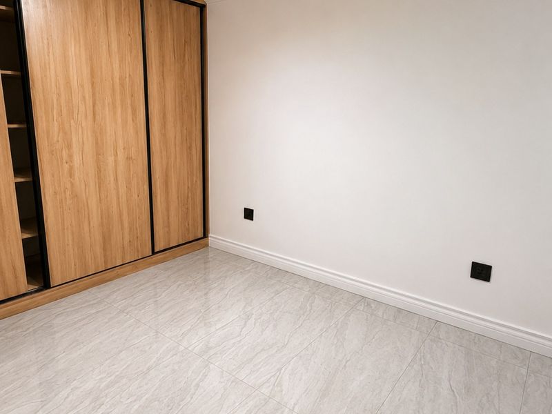 A clean, modern bedroom featuring light-toned tiled flooring, a large built-in wooden wardrobe with sliding doors, and contemporary track lighting.