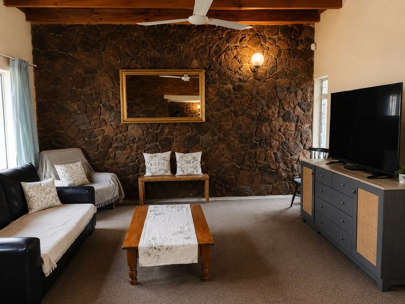 A cozy living room featuring a prominent stone feature wall, exposed timber ceiling beams, and a ceiling fan, creating a rustic and inviting atmosphere.