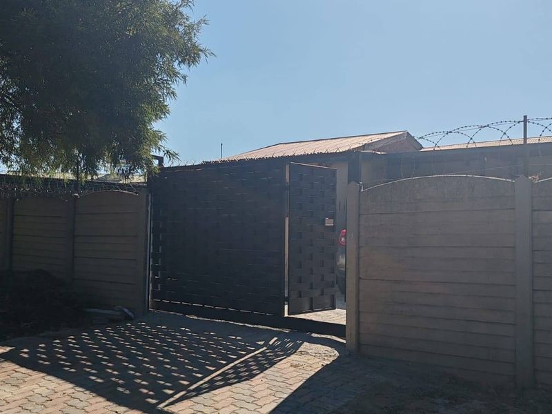 Front exterior view featuring a paved driveway, metal gate, and precast concrete boundary wall topped with razor wire security fencing.