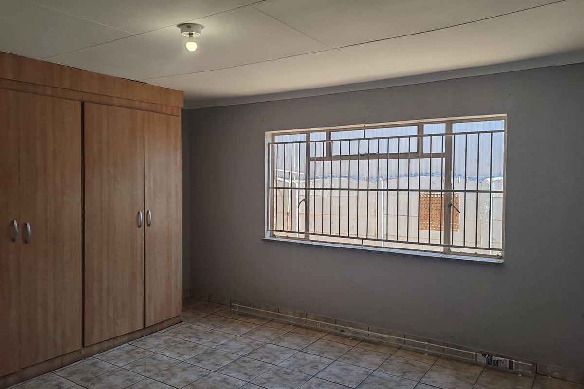 A vacant bedroom featuring tiled flooring, built-in wooden cupboards, and a window fitted with security bars.