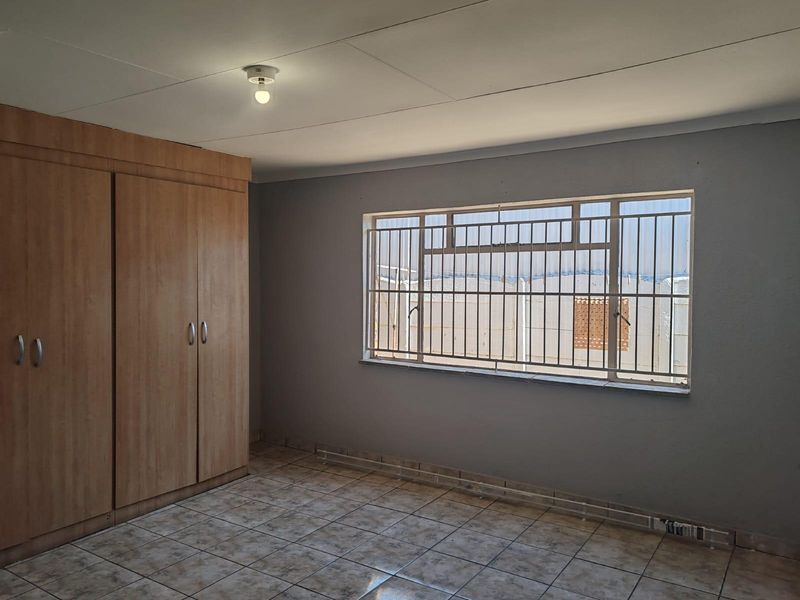 A vacant bedroom featuring tiled flooring, built-in wooden cupboards, and a window fitted with security bars.