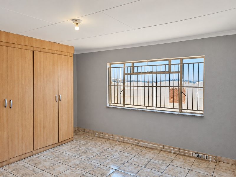 A clean, empty bedroom featuring built-in wooden cupboards, tiled flooring, and a window fitted with security bars.