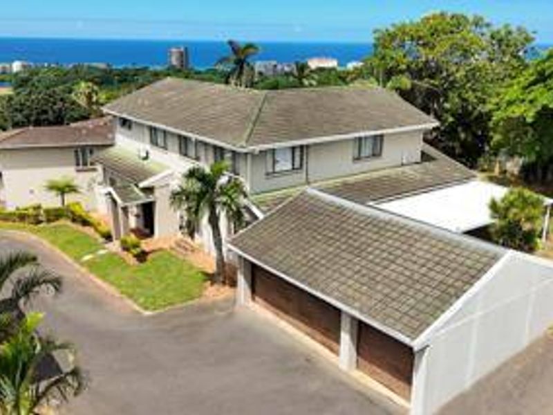 Aerial view of a townhouse property featuring a tiled roof, double garage, paved driveway, and a covered patio with scenic coastal views.
