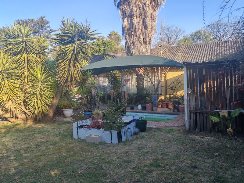 A backyard pool area featuring a small swimming pool with a shade sail, surrounded by established garden landscaping and a paved patio area.