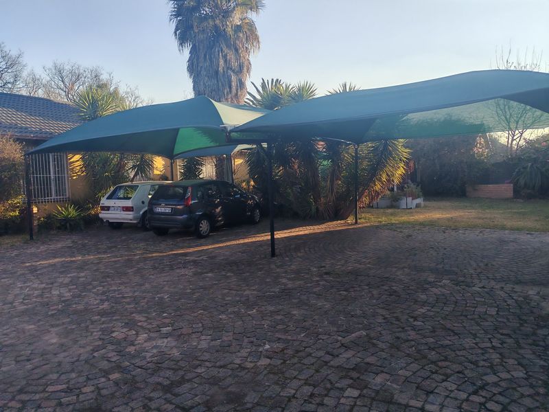 Front exterior view featuring a large shade cloth carport structure over a cobblestone driveway with established garden landscaping.