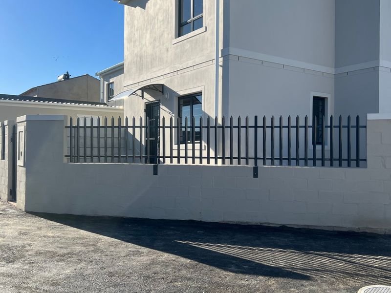 Front elevation of a modern two-story house featuring a rendered facade, metal security fencing, and a paved driveway area.
