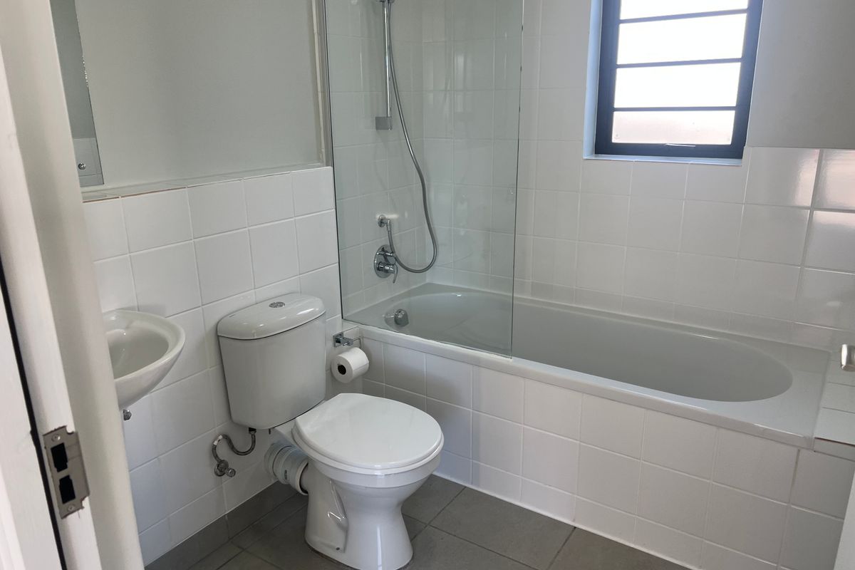 A clean, functional bathroom featuring a built-in bathtub with a glass shower screen, wall-mounted basin, and neutral ceramic tiling throughout.