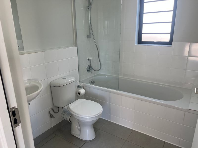 A clean, functional bathroom featuring a built-in bathtub with a glass shower screen, wall-mounted basin, and neutral ceramic tiling throughout.