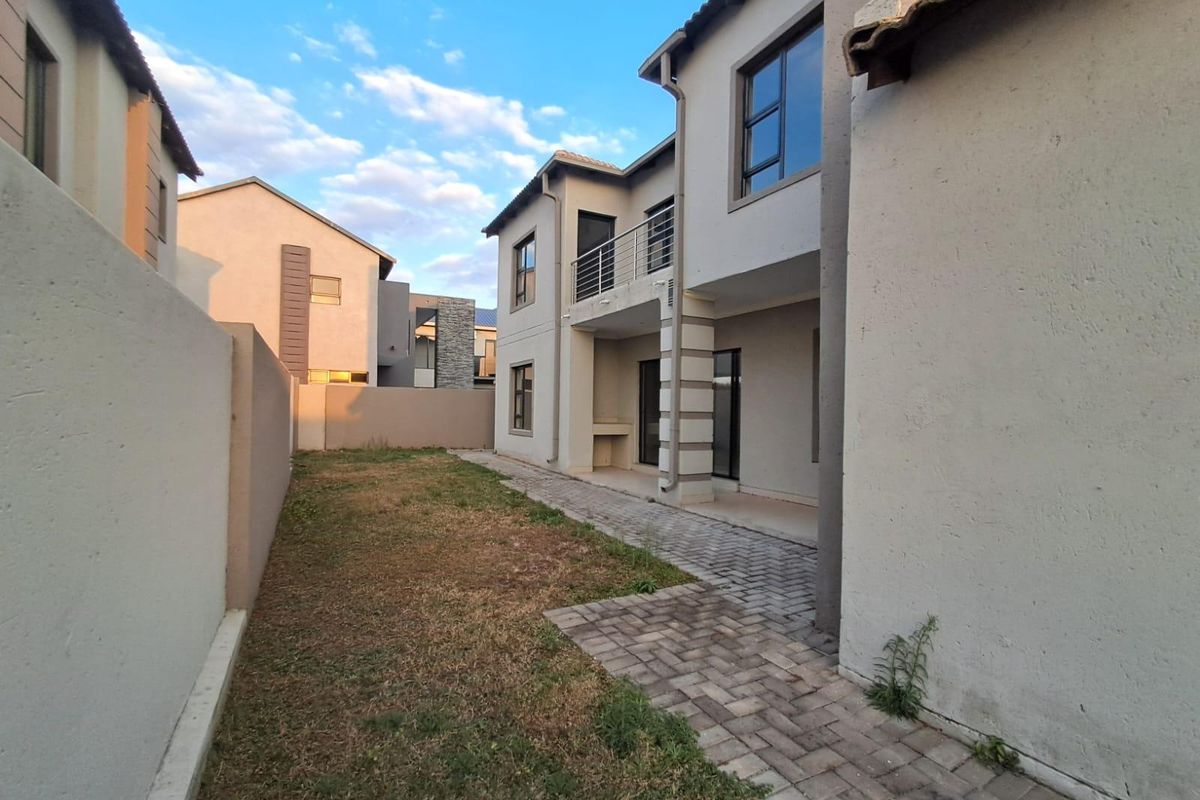 Rear view of the property featuring a paved walkway, a small lawn area, a built-in braai, and an upper-level balcony.
