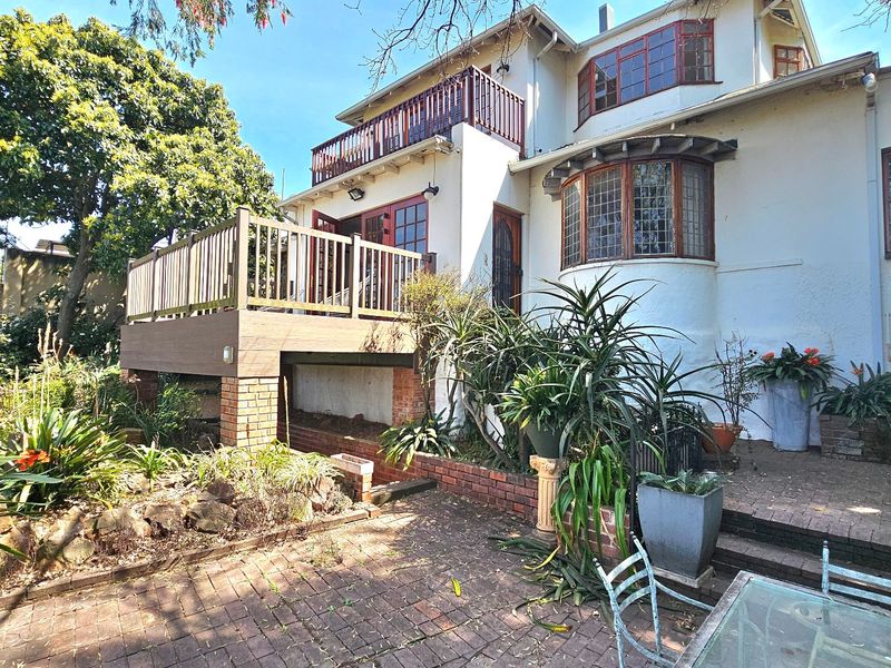 Rear view of the property featuring a raised timber deck, balcony, and brick paved patio area surrounded by established garden landscaping.