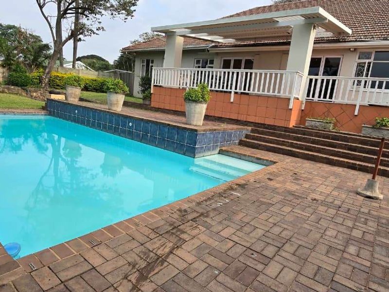 A spacious outdoor pool area featuring a swimming pool, paved patio, and a covered pergola attached to the rear of the house.