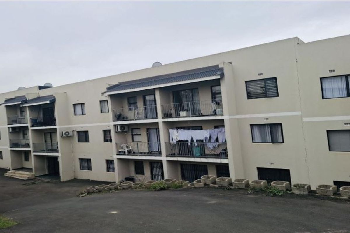 Rear view of a multi-story apartment complex featuring private balconies, metal railings, and an asphalt driveway with concrete retaining wall elements.
