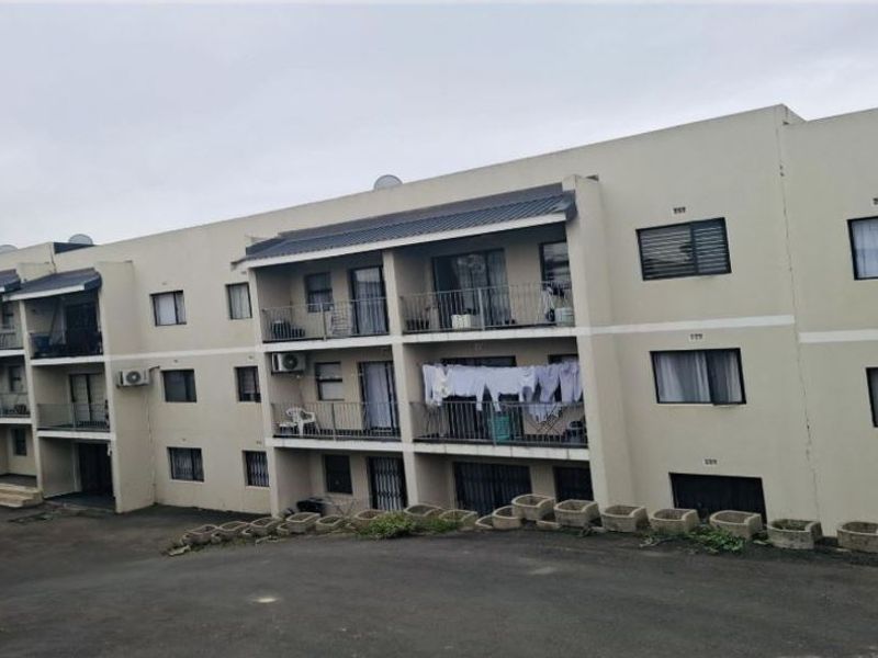 Rear view of a multi-story apartment complex featuring private balconies, metal railings, and an asphalt driveway with concrete retaining wall elements.