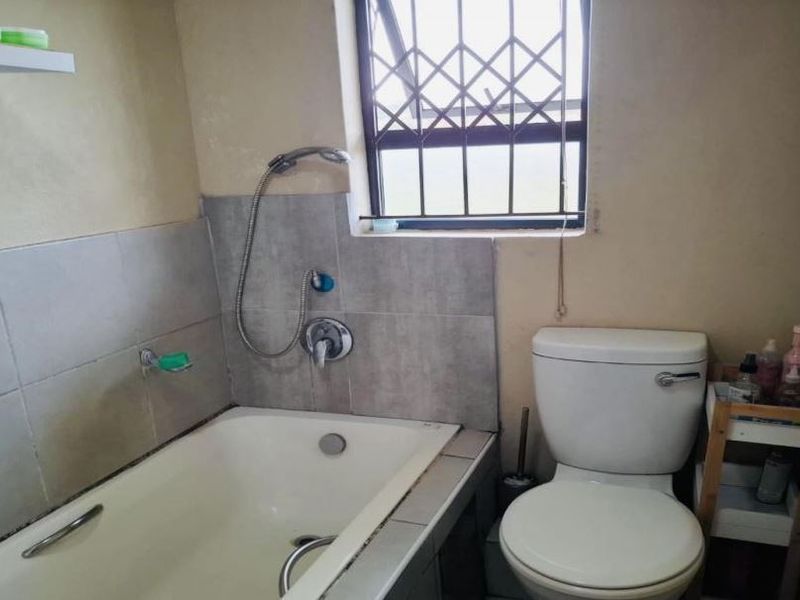 A compact bathroom featuring a built-in bathtub with shower mixer, a standard toilet, and a window fitted with a security gate.