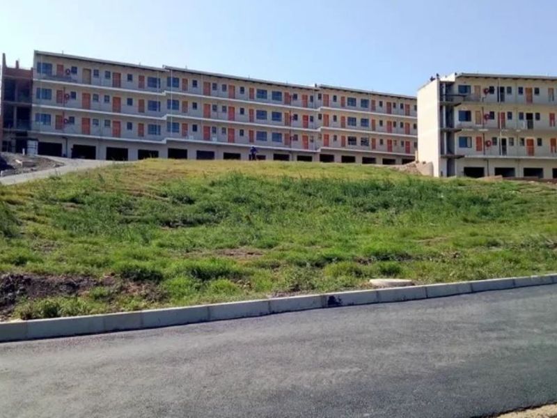 Exterior view of a multi-story apartment complex under construction, featuring paved road access and an unfinished landscaped embankment.