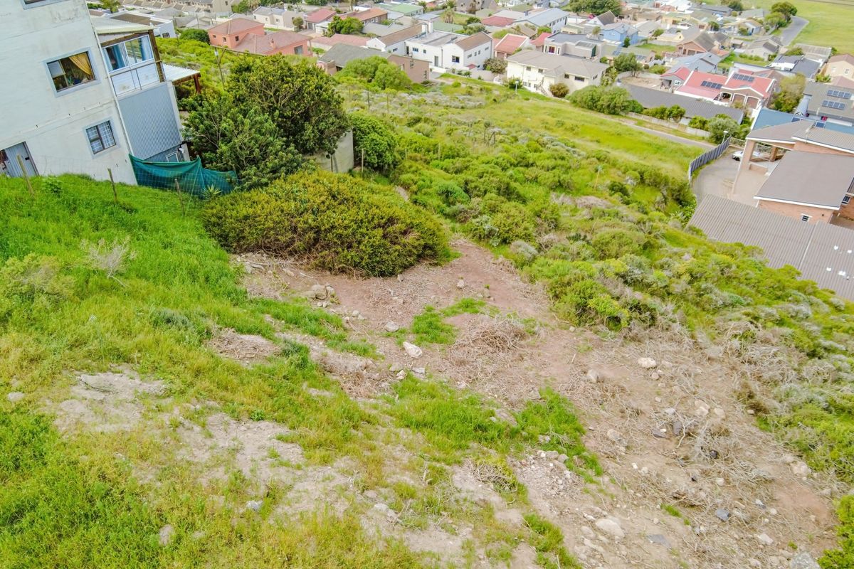 An aerial view of a sloped, undeveloped residential plot with natural vegetation, situated in a suburban neighborhood.
