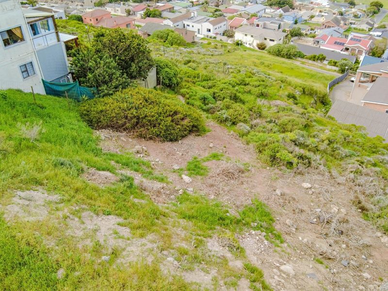 An aerial view of a sloped, undeveloped residential plot with natural vegetation, situated in a suburban neighborhood.