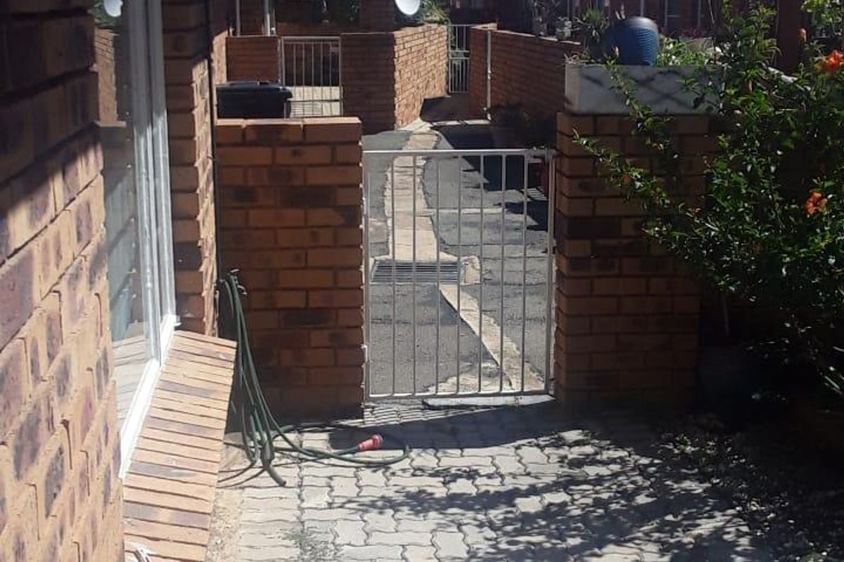 A view of the rear exterior area featuring brick paving, a secure metal gate, and brick boundary walls within a townhouse complex.
