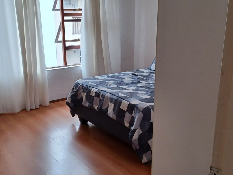 A bedroom featuring laminate flooring and a large window, providing natural light and a view of the surrounding building exterior.