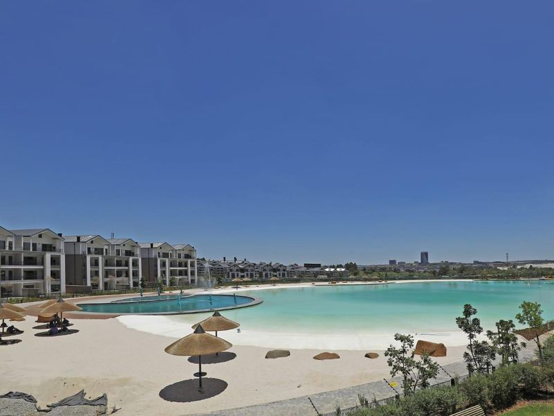 A wide-angle view of a large communal swimming pool and man-made beach area within a modern apartment complex.
