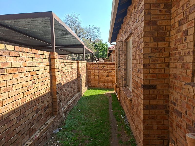 A side exterior view of the townhouse featuring face brick walls, a metal carport structure, and a narrow grassed pathway.