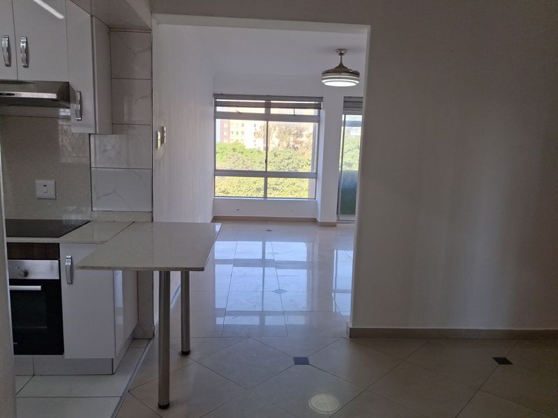 An open-plan kitchen and living area featuring tiled flooring, a built-in oven, and a breakfast counter, leading to a bright windowed living space.