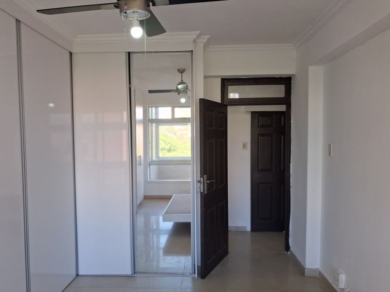 A bright, empty bedroom featuring modern white built-in cupboards with mirrored doors, tiled flooring, and a ceiling fan.