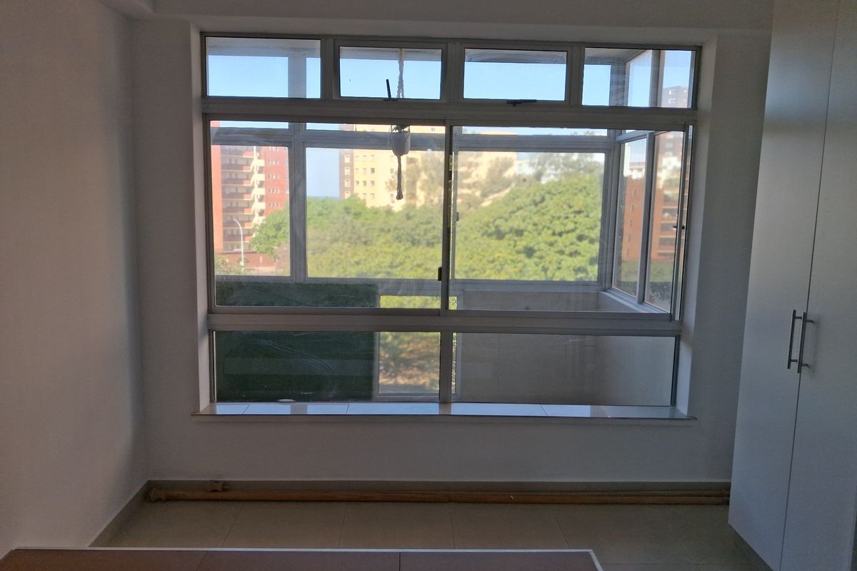 A bright, empty bedroom featuring tiled flooring, a built-in wardrobe, and large aluminium-framed windows offering an elevated view of the surrounding neighbourhood.