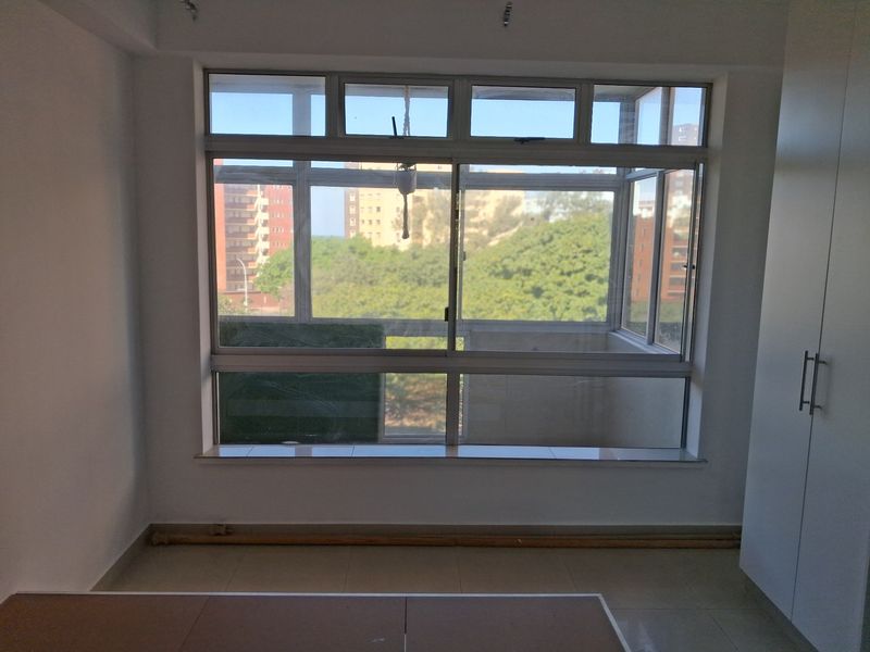 A bright, empty bedroom featuring tiled flooring, a built-in wardrobe, and large aluminium-framed windows offering an elevated view of the surrounding neighbourhood.