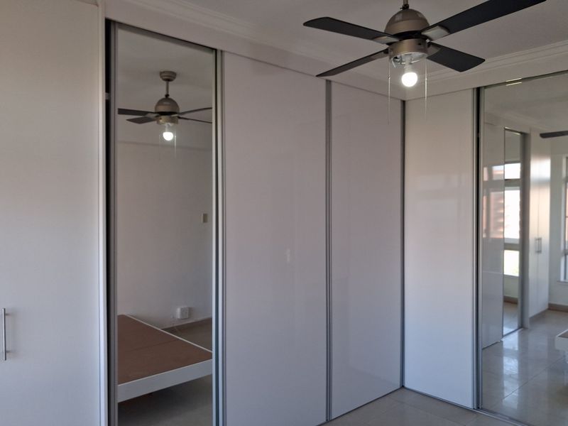 A clean bedroom featuring modern built-in cupboards with mirrored sliding doors, a ceiling fan, and neutral tiled flooring.