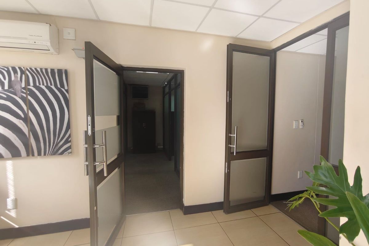 A bright office space featuring tiled flooring, suspended ceilings, and modern aluminium-framed glass doors leading to an adjacent carpeted corridor.