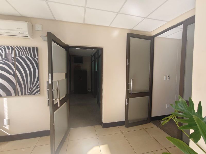A bright office space featuring tiled flooring, suspended ceilings, and modern aluminium-framed glass doors leading to an adjacent carpeted corridor.