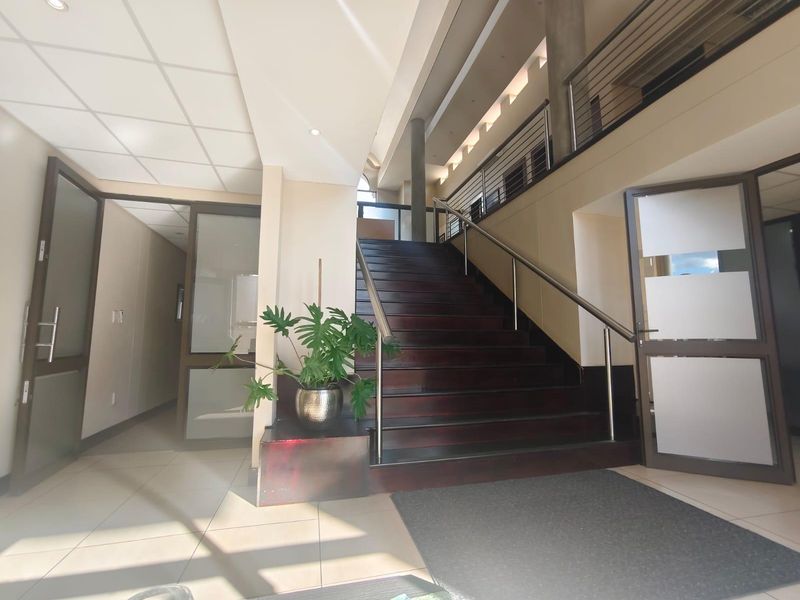 A bright and spacious commercial entrance foyer featuring a prominent wooden staircase with stainless steel handrails, tiled flooring, and access to multiple office areas.