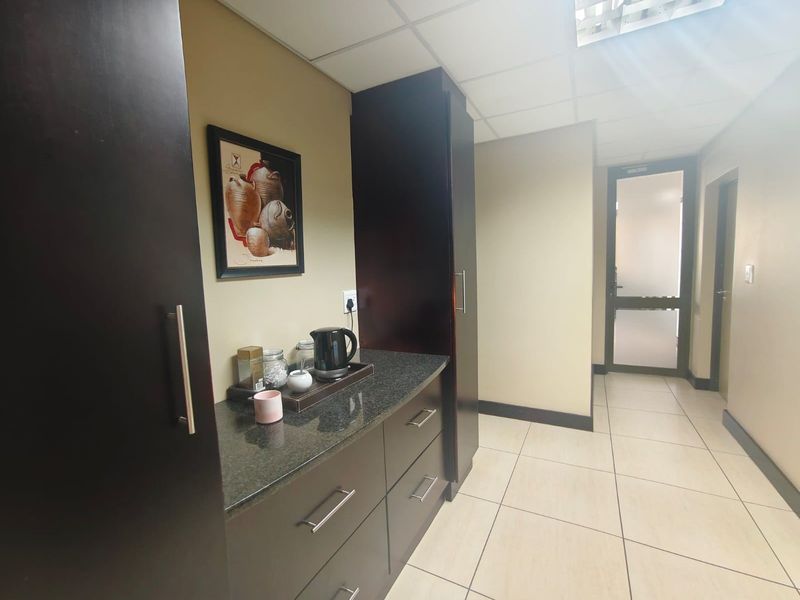 A compact kitchenette area featuring dark wood cabinetry, granite countertops, and tiled flooring, leading into a hallway with a glass-paneled door.